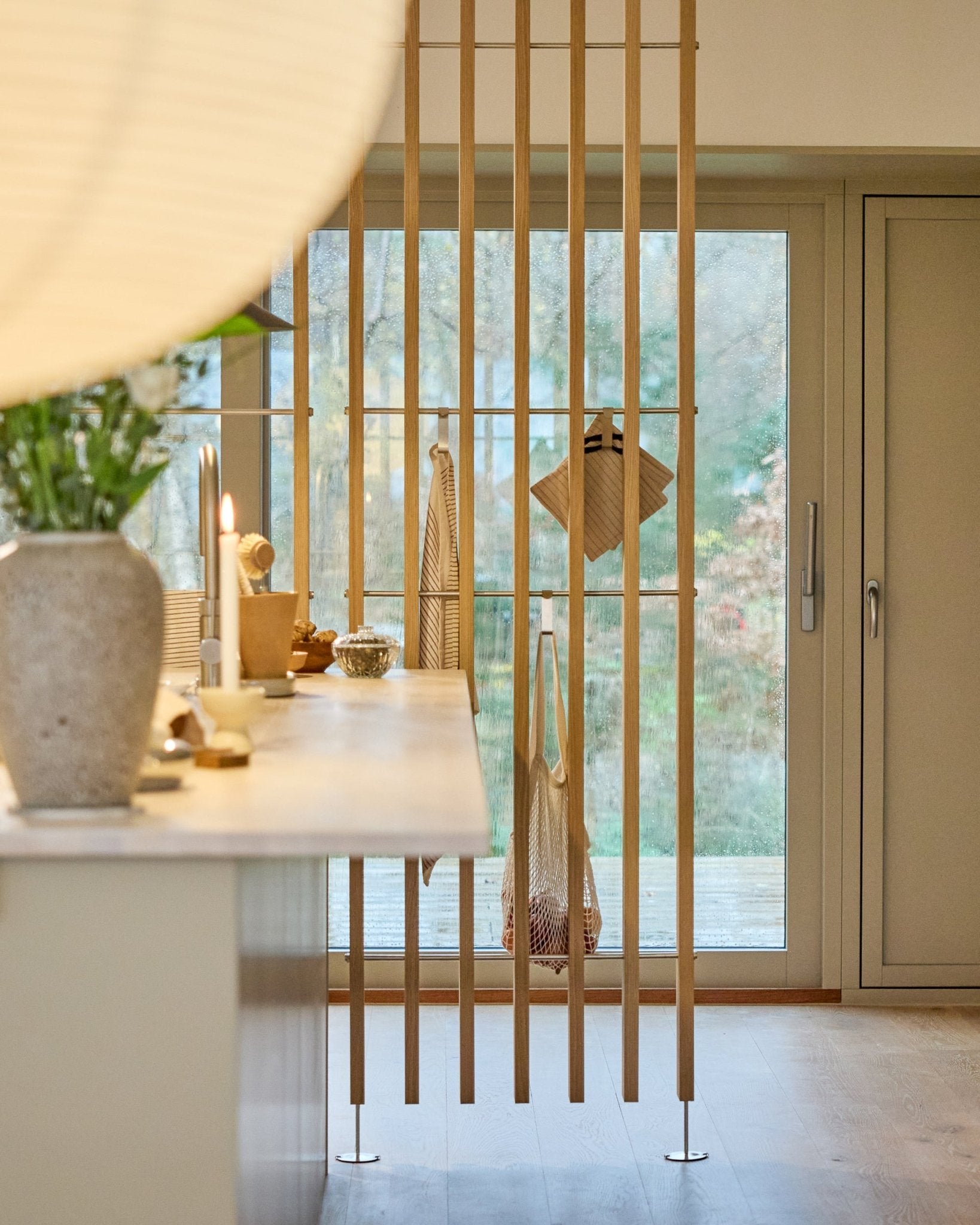 Minimalist kitchen featuring a WoodUpp Embrace room divider (BM1, 286-302 cm) in oak veneer, with light countertops, floral vase, and garden views.